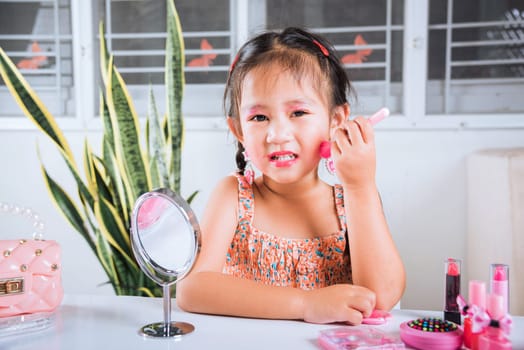 Asian adorable funny little girl making makeup her face she look in the mirror and cheek fluffy brush for powder, Learning activity to be woman, happy kid is beautiful make up with cosmetics toy