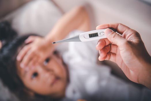Sick kid. Mother checking temperature of her sick daughter with thermometer in mouth, child laying in bed taking measuring her temperature for fever and illness