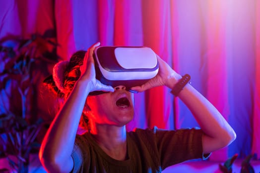 Asian woman wear the VR helmet excited emotional at home purple and blue background, female experiencing virtual reality goggles experiencing reality, metaverse and cyberspace concept