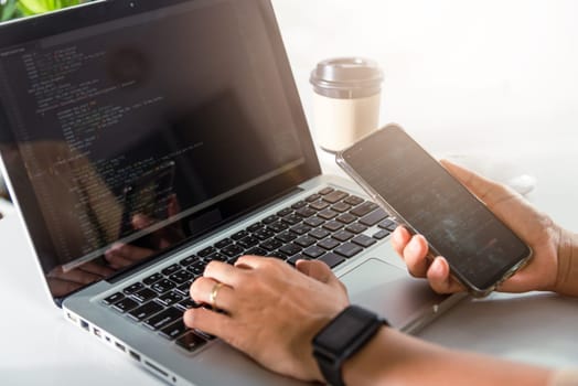Programmer woman looking mobile phone program code at workplace, software developer hands typing data coding programming javascript with laptop computer, IT startup concept, Web application design