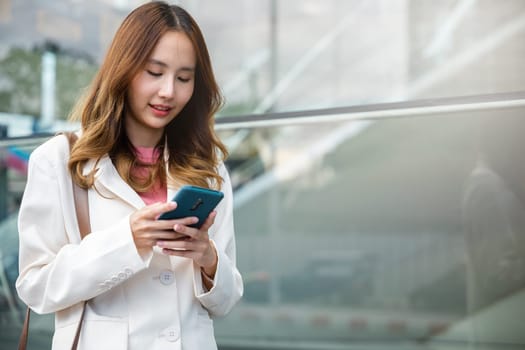 Asian businesswoman typing text on mobile phone standing outdoors street front building near office, Portrait successful business woman smiling holding smartphone using app chat online in morning