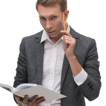 A man in a grey suit holds a book while pondering with a pencil in hand, appearing deep in thought against a plain background.