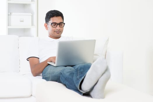 Young Indian guy doing online business with laptop computer at home. Asian man using internet indoor, relaxed and sitting on sofa. Handsome male model.