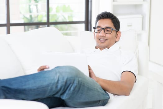 Indian guy using digital computer tablet at home. Asian man relaxed and lying on sofa indoor. Handsome male model.