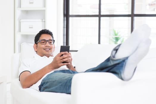 Smartphone mobile apps concept. Indian guy using smart phone. Asian man relaxed and lying on sofa indoor. Handsome male model.