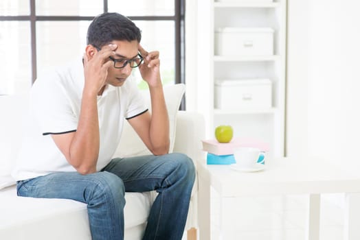 Indian guy having headache at home. Asian man hands holding head sitting on sofa indoor. Handsome male portrait.