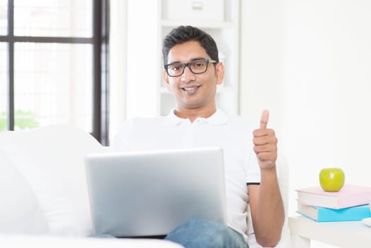 Indian guy with laptop computer working from home and giving thumb up. Asian man using internet indoor, relaxed and sitting on sofa. Handsome male model.