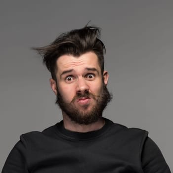 Portrait of young tousled man with a beard and mustache in studio  on gray background. Concept of confusion and dissatisfaction