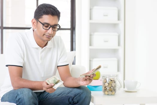 Too less money. Indian guy counting money with sad expression, sitting on sofa at home. Asian male indoor portrait.