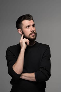 Fashion portrait of young  thoughtful  man in black  poses over gray background. hand man scratching his beard. gaze directed upwards