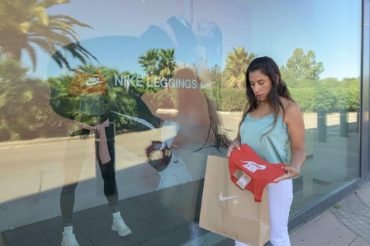 mallorca, balearic islands spain-november 05 2022: hispanic woman looking at the window of the nike sports store happy comparing the purchase made in the mall mallorca fashion outlet, on the outskirts of the city of palma