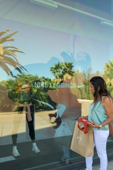 mallorca, balearic islands spain-november 05 2022: hispanic woman looking at the window of the nike sports store happy comparing the purchase made in the mall mallorca fashion outlet, on the outskirts of the city of palma