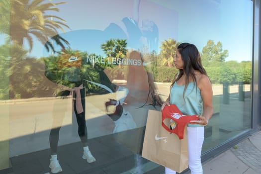 mallorca, balearic islands spain-november 05 2022: hispanic woman looking at the window of the nike sports store happy comparing the purchase made in the mall mallorca fashion outlet, on the outskirts of the city of palma