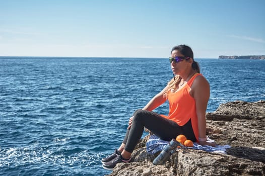 Latin woman, middle-aged, resting, regaining strength, eating, drinking water, after a gym session, wearing orange top, burning calories, keeping fit, outdoors by the sea, blue sky, beautiful day , sunny winter, Bluetooth headphones, and smart watch