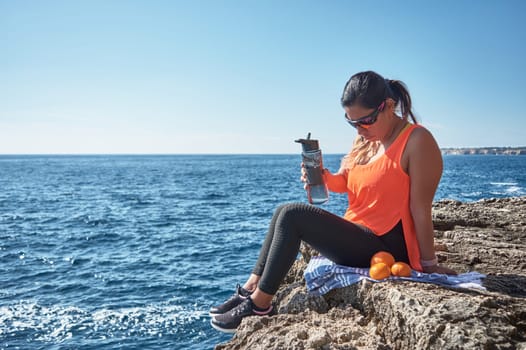 Latin woman, middle-aged, resting, regaining strength, eating, drinking water, after a gym session, burning calories, keeping fit, outdoors by the sea, wearing headphones and smart watch