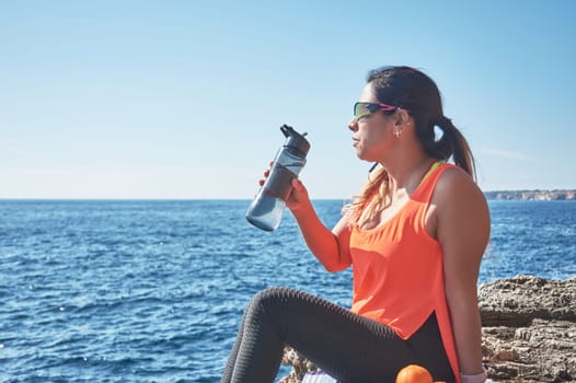 Latin woman, middle-aged, resting, regaining strength, eating, drinking water, after a gym session, burning calories, keeping fit, outdoors by the sea, wearing headphones and smart watch