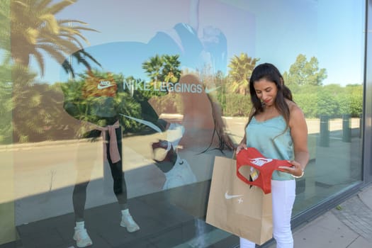mallorca, balearic islands spain-november 05 2022: hispanic woman looking at the window of the nike sports store happy comparing the purchase made in the mall mallorca fashion outlet, on the outskirts of the city of palma