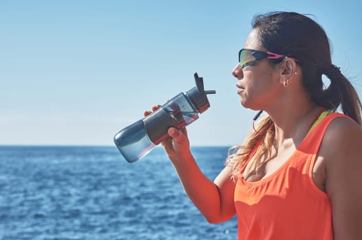 Latin woman, middle-aged, resting, regaining strength, eating, drinking water, after a gym session, burning calories, keeping fit, outdoors by the sea, wearing headphones and smart watch