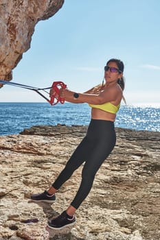 FITNESS LATIN WOMAN IN SPORTS SET TRAINING WITH ELASTIC BAND, WEIGHTS, GYM EXERCISES, IN FRONT OF THE WATER.