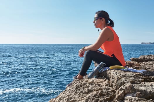 Latin woman, middle-aged, resting, regaining strength, eating, drinking water, after a gym session, burning calories, keeping fit, outdoors by the sea, wearing headphones and smart watch