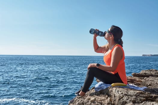 Latin woman, middle-aged, resting, regaining strength, eating, drinking water, after a gym session, burning calories, keeping fit, outdoors by the sea, wearing headphones and smart watch