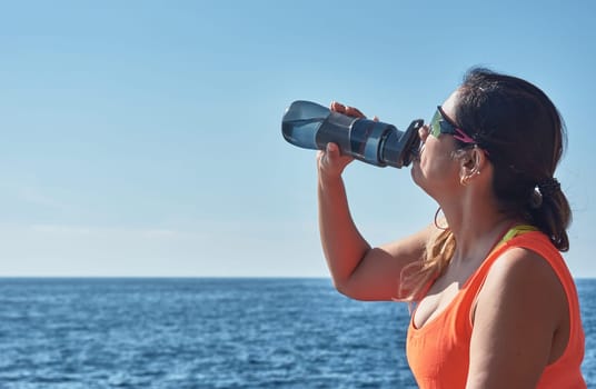 Latin woman, middle-aged, resting, regaining strength, eating, drinking water, after a gym session, wearing orange top, burning calories, keeping fit, outdoors by the sea, blue sky, beautiful day , sunny winter, Bluetooth headphones, and smart watch