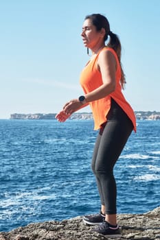 FITNESS LATIN WOMAN IN SPORTS SET TRAINING WITH ELASTIC BAND, WEIGHTS, GYM EXERCISES, IN FRONT OF THE WATER.