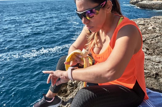 Latin woman, middle-aged, resting, regaining strength, eating, drinking water, after a gym session, burning calories, keeping fit, outdoors by the sea, wearing headphones and smart watch
