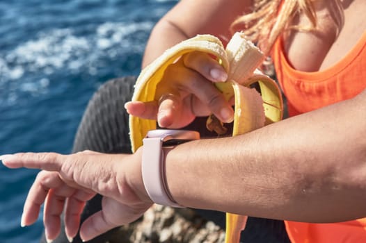 Latin woman, middle-aged, resting, regaining strength, eating, drinking water, after a gym session, burning calories, keeping fit, outdoors by the sea, wearing headphones and smart watch
