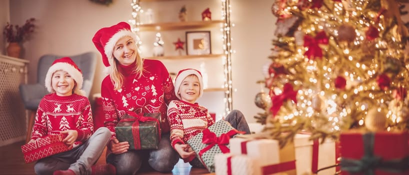 Mother and her son ad daughter sitting indoors with Christmas present boxes in their hands. Concept of celebrating of Christmastime and New Year together.