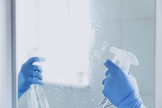 Cropped view of housekeeper woman hand in rubber blue gloves spraying chemical cleaner liquid to the mirror while cleaning bathroom at home. Housekeeping and cleaning service. Clean house, cleanliness