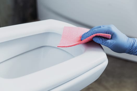 Woman hand closeup gloved in rubber protective gloves. Housewife cleaning toilet bowl, seat with detergent, pink cloth in bathroom or public restroom. Disinfection, hygiene, cleaning service concept