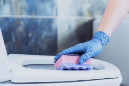 Cropped image view with close up on woman hands gloved in rubber protective gloves. Housewife cleaning toilet bowl, seat with pink sponge in bathroom or modern public restroom with blue grey tile