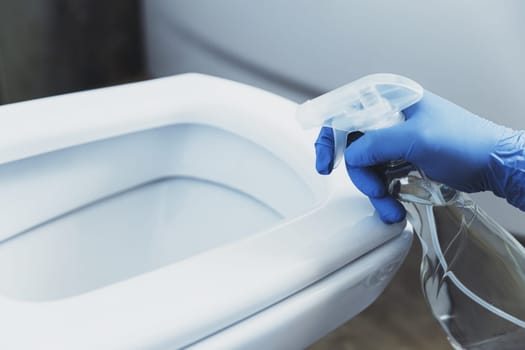 Close up on woman hand gloved in blue rubber protective gloves. Housewife cleaning toilet bowl, seat with detergent liquid in bathroom or restroom. Disinfection, hygiene, cleaning service concept