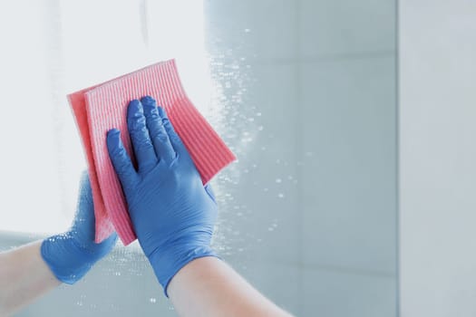 Cropped view of housekeeper woman hand in blue rubber glove polishing, cleaning, swiping mirror with cloth rag, spraying cleaner liquid while cleaning bathroom at home. Housekeeping, cleaning service