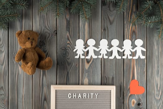 Composition of teddy bear toy and paper children garland cutting from paper near nameplate charity on wooden Christmas background. Charity for child care, orphan kids. Flat lay, top view, copy space