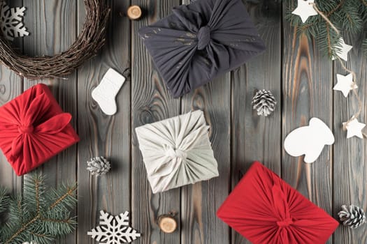 Christmas festive holiday composition of red, white, blue Furoshiki present boxes with decorated spruce branches, wreath, festive socks, mitten, star, cone garland on wooden background. Flat lay