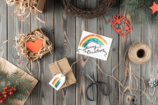Composition of present box in packaging paper, box with heart and rainbow label, festive card merry Christmas with scissors, rope on wooden table with fir twigs, wreath, cone decoration. Flat lay