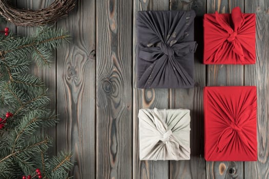 Holiday composition four handmade, craft, wrapped Furoshiki present boxes with decorated fir tree branches and wreath on dark wooden background. Holiday tradition for Christmas. Flat lay, copy space