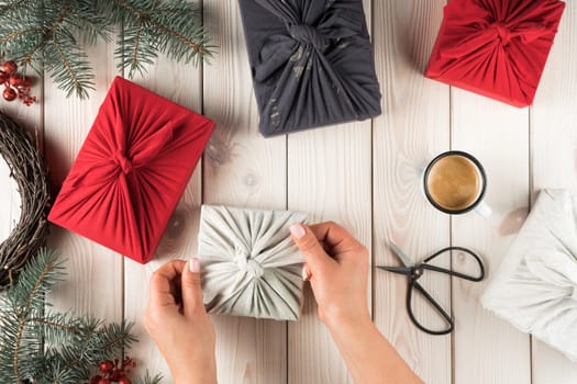 Unrecognizable woman hands making ecological, natural furoshiki presents on coffee break for holidays on wooden decorative background. Do it yourself, tutorial of craft Christmas gifts. Top view
