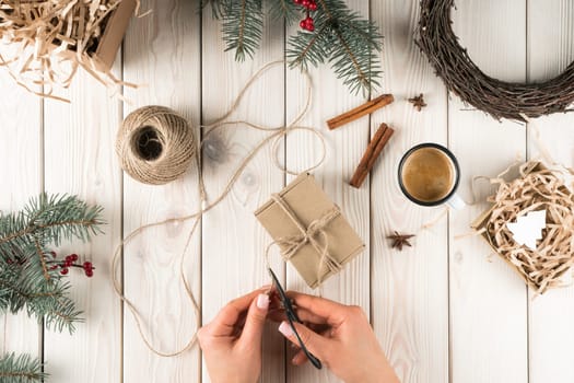 Cropped woman hands cutting rope for crafted cardboard box in xmas decoration of cinnamon, gifts and fir tree branch and wreath. Natural wrapping and packaging of present boxes for shipping. Top view