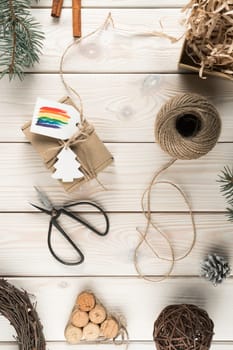 Vertical composition of natural, organic, fabric wrapped box with rainbow label, festive cards with scissors and rope sleave on wooden table with holiday fir tree, wreath brunches decoration. Flat lay