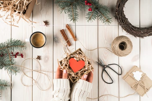 Cropped woman hands in white sweater holding open cardboard box with red heart in xmas decoration of cinnamon, scissors, presents and fir tree twigs and wreath. Handmade crafted gifts for holidays
