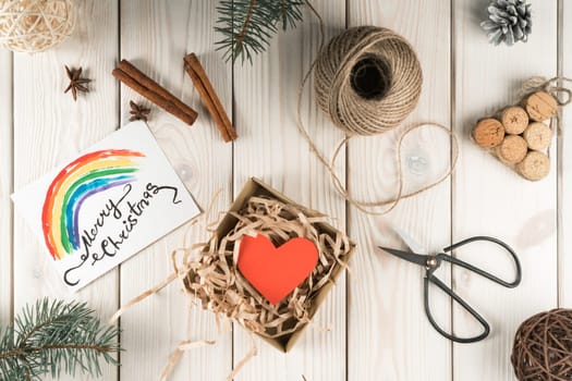 Open fabric present box with red heart and rainbow label card merry christmas. DIY, handmade gifts with scissors and wrapping paper with fir, nuts and cinnamon decorations on wooden background
