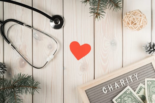 Abstract composition of nameplate charity with money, stethoscope, red heart and spruce twigs as decoration on wooden background. Healthcare and medicine, charity, donation. World health day. Top view