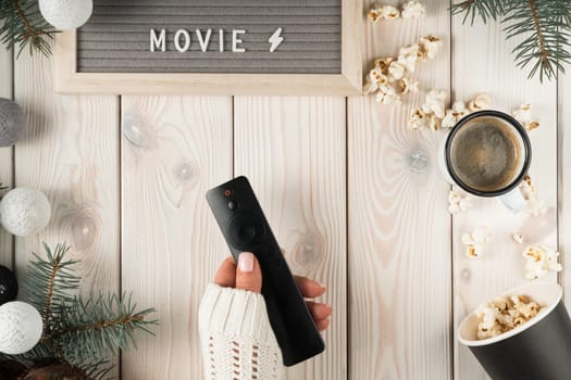 Cropped woman hands in cozy sweater holding remote control, nameplate movie, cup of coffee, bucket of popcorn on decorative xmas wooden background. Weekend activity with tv set, relax. Top view