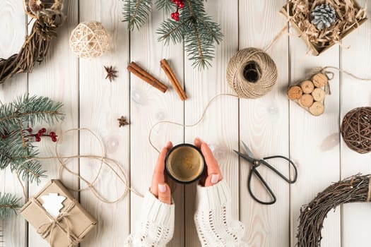 Unrecognizable woman hands in warm sweater hold hot cup of coffee in xmas decoration of cinnamon, scissors, fir twigs, nuts and wreath. Christmas Eve design and handicraft present at home. Top view