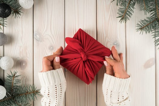 Cropped woman hands in warm sweater tying bows on red furoshiki present box with spruce twigs and Christmas toys decoration on wooden background. Ecological, natural holiday gift. Copy space, top view