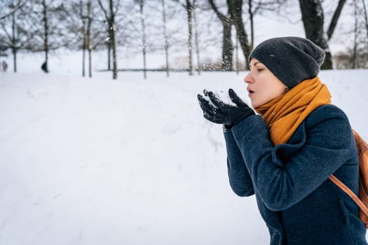 A woman in winter clothes blows snow off her palms in gloves.