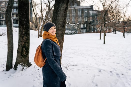A woman in winter clothes with a leather red backpack looks around smiling.
